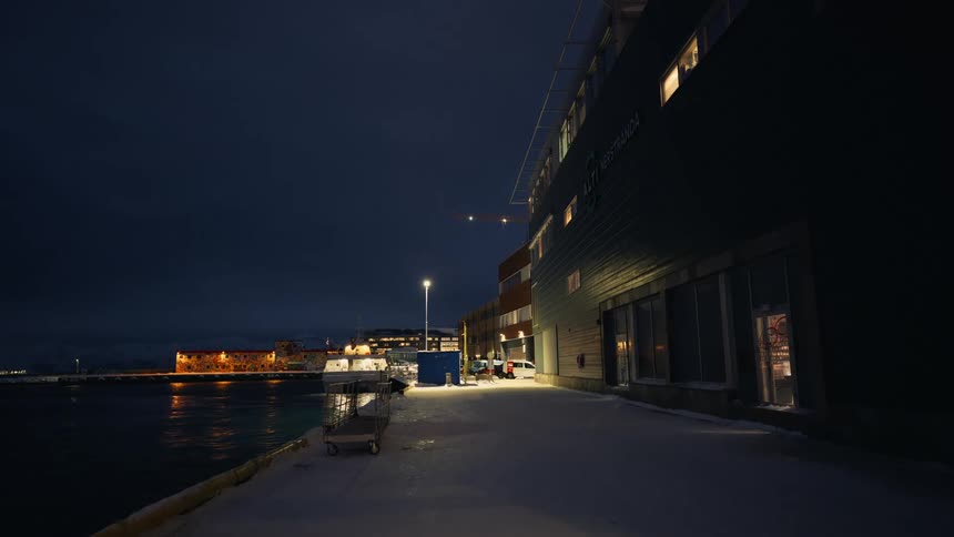 A small boat slowly cruises through a tranquil, snow-covered Norwegian harbor at night