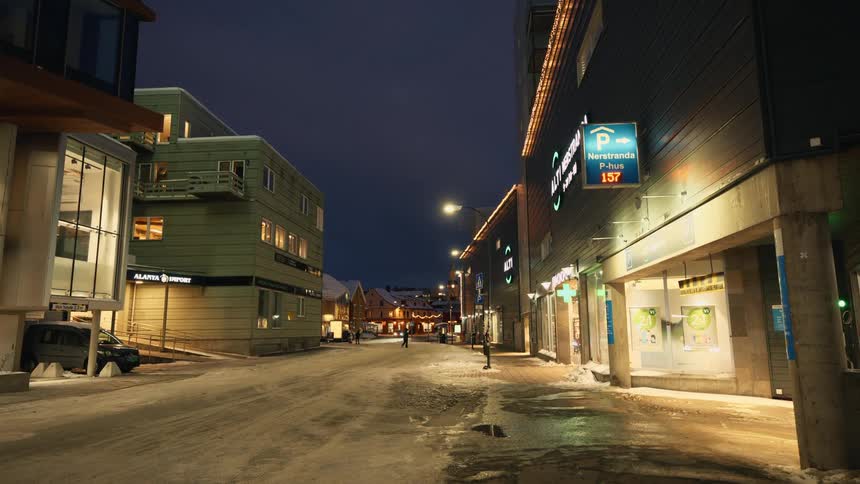 A quiet snow-covered street in a modern Norwegian town is illuminated by warm streetlights at night