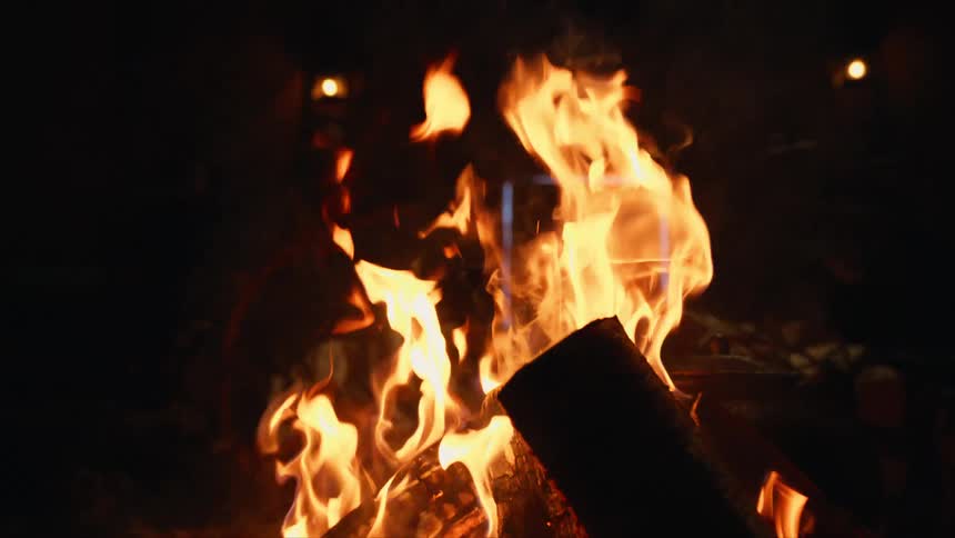 A warm campfire burns in slow motion at night with a laptop glowing in the background