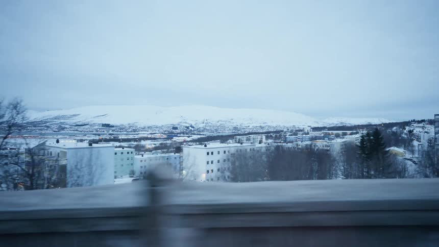 A slow-motion view from a moving vehicle reveals a snowy town and distant mountains in a cold, northern landscape