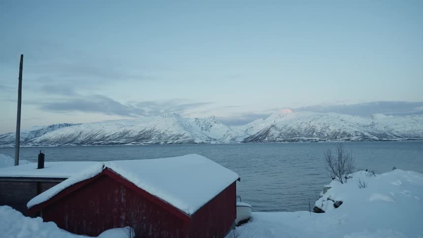 A tranquil winter scene shows a snow-covered red shed on the banks of a calm fjord with majestic mountains in the distance