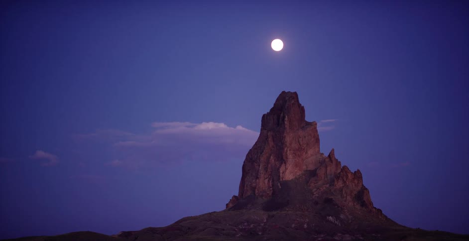 A bright full moon hangs in the deep blue twilight sky over the iconic Agathla Peak rock formation in the Arizona desert