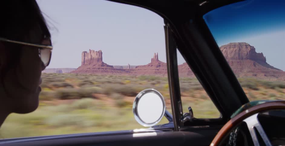 Over-the-shoulder view of a woman in sunglasses driving a classic car through the desert with Monument Valley in the background