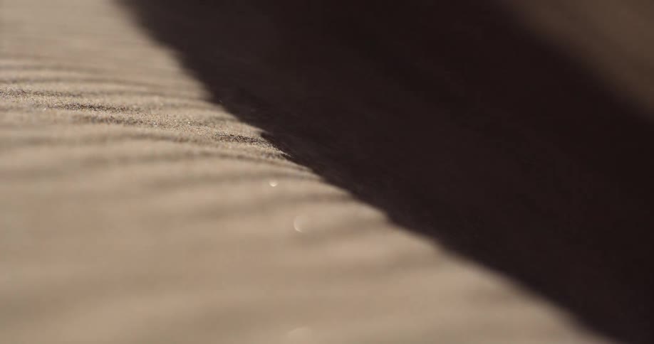 A mesmerizing close-up, slow-motion shot of wind blowing fine sand grains over the shadowed ridge of a desert dune