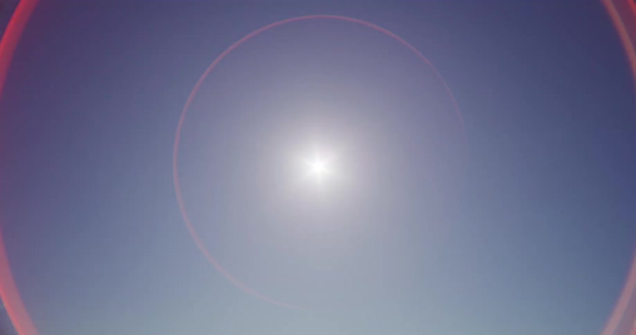A bright sun shines in a clear, cloudless sky, creating a prominent red circular lens flare