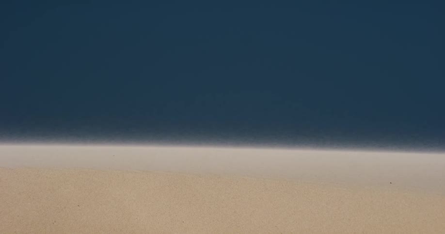 A minimalist shot of fine sand blowing across a dune against a solid dark blue background