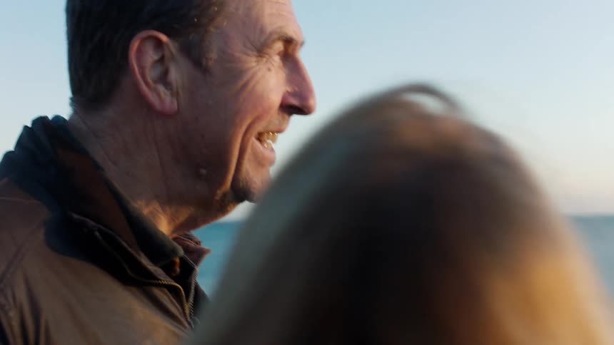 A close-up slow-motion shot captures a mature man smiling lovingly at his partner as they enjoy the ocean view at sunset