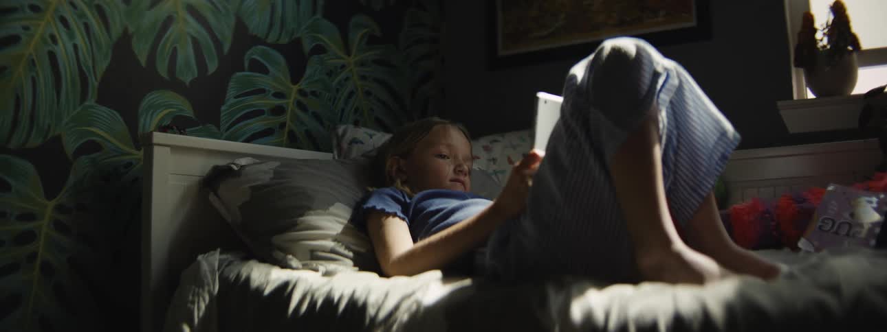Sunlight streams into a dark bedroom as a young girl in pajamas lies in bed watching a digital tablet