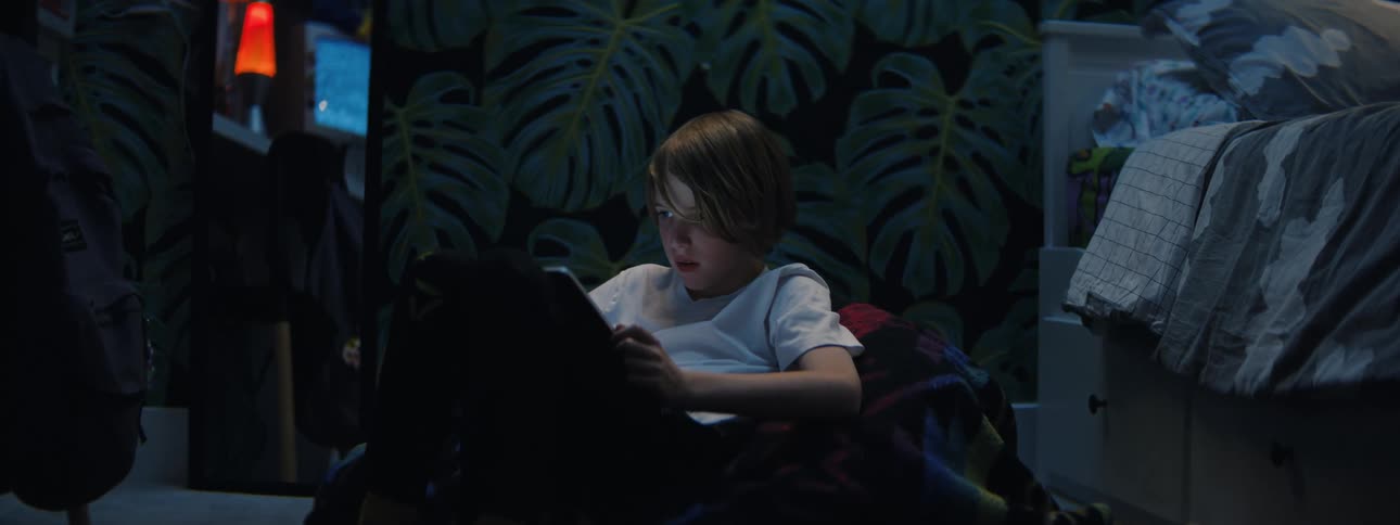 A young boy sits on the floor of his dimly lit bedroom at night, intently focused on his digital tablet