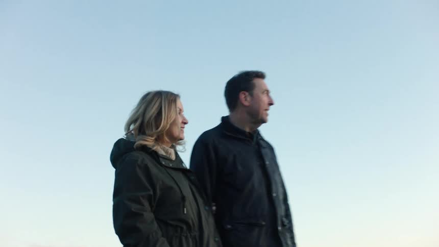 An expecting couple in warm jackets stands on a grassy cliff, smiling and pointing out at the serene ocean view together