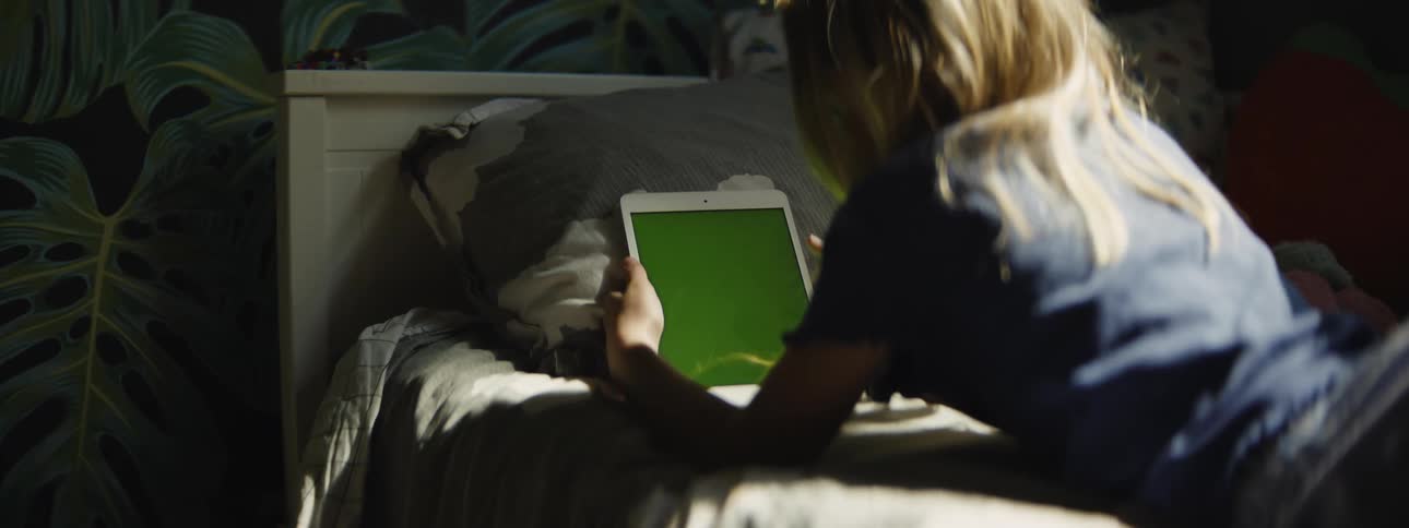 A young girl lies on her stomach in a sun-drenched bedroom, holding a tablet with a green screen