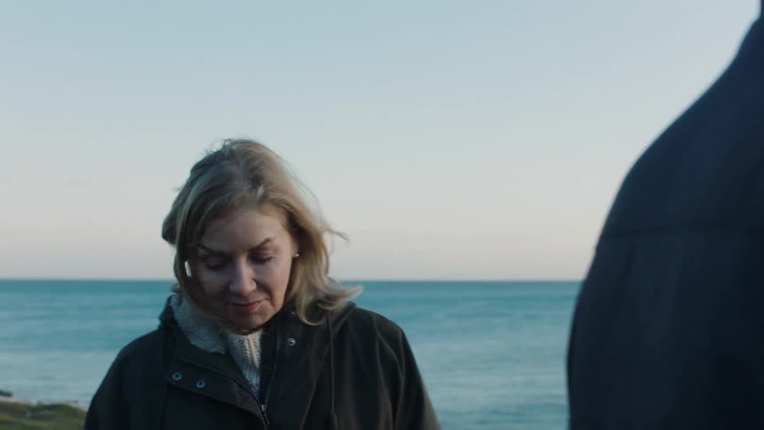 A close-up shot of a thoughtful woman talking with a companion by the sea as the wind blows her hair