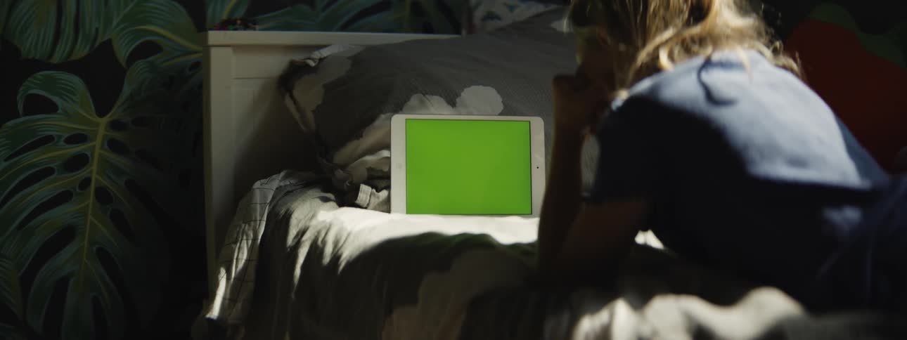 A young person lies on their bed in a sunlit room watching a tablet with a green screen as the camera slowly zooms in