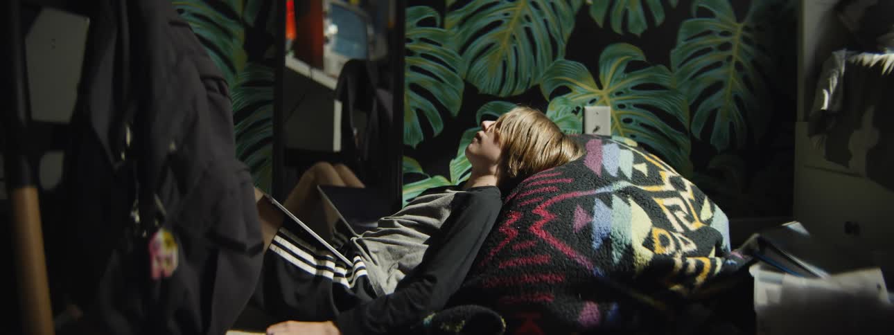 A tired young person using their laptop falls asleep on the floor of their sunlit, leaf-patterned bedroom