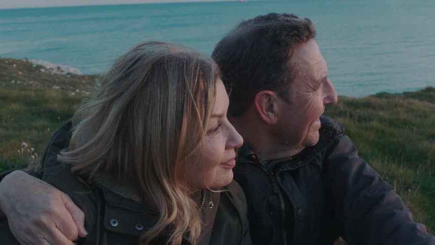 A content middle-aged couple enjoys a peaceful moment together, sitting on a grassy clifftop and looking out at the ocean at dusk