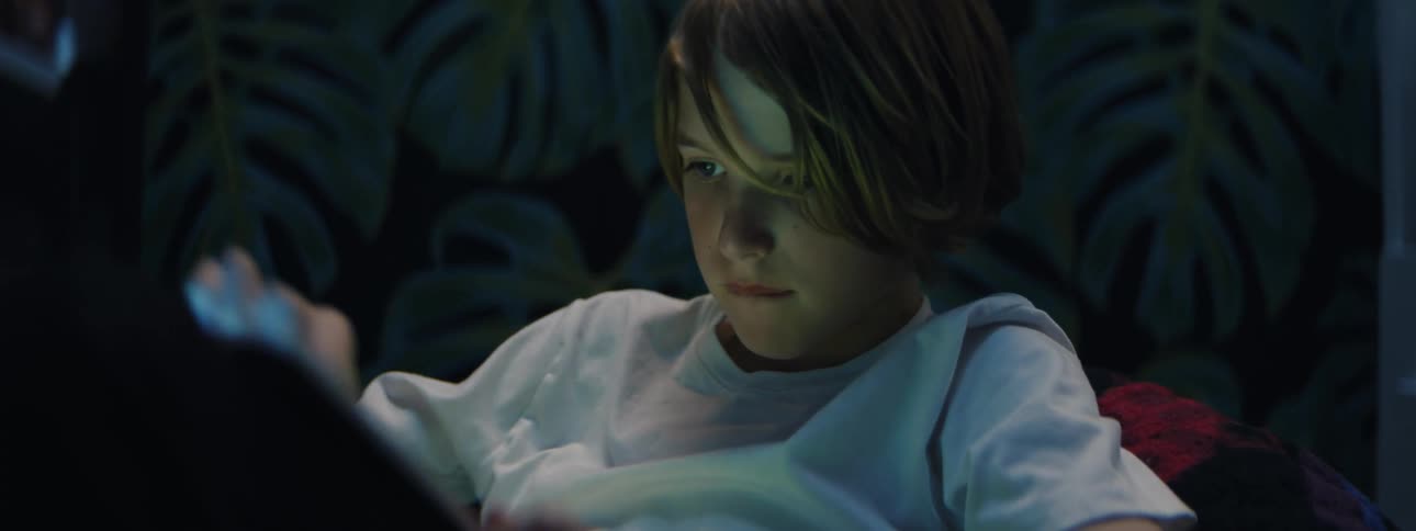 Boy sits in bed at night absorbed in a tablet screen in a dimly lit bedroom