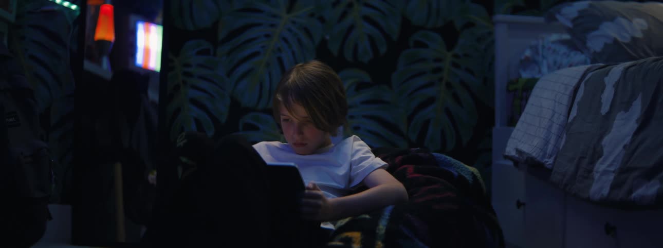 A young boy sits on the floor of his dimly lit bedroom at night, intensely focused on the glowing screen of his tablet