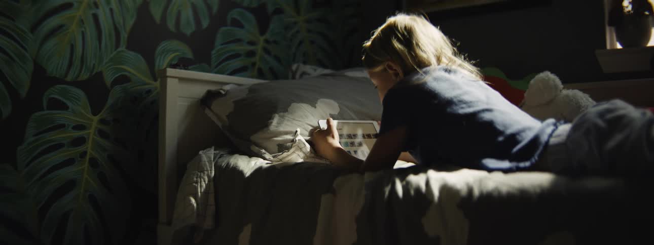 Young girl lies on her bed in a dimly lit room, engrossed in playing a game on her tablet as sunlight streams in from a window