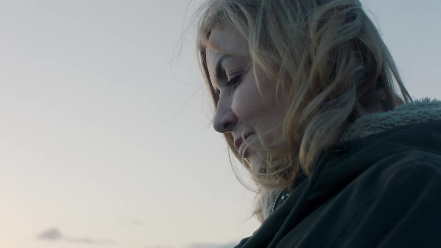 A thoughtful woman in profile holds a delicate flower while watching the sunset over the ocean