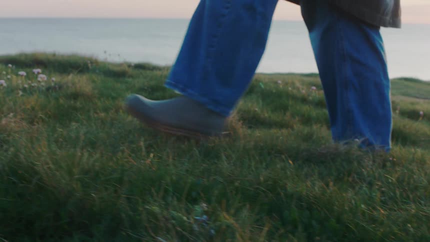 A person wearing jeans and boots walks in slow motion along a grassy coastal cliff with the ocean in the background at sunrise