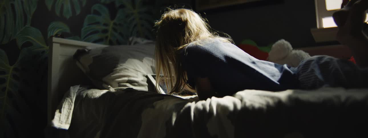 A young girl bathed in a single ray of sunlight lies on her stomach in bed, engrossed in using a digital tablet