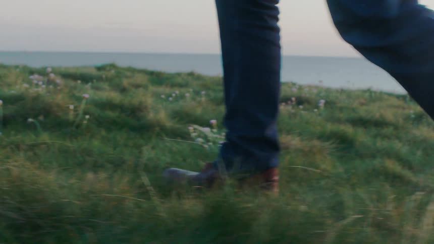 Close-up slow motion shot of a person's feet in leather boots walking through a grassy field on a cliff overlooking the sea