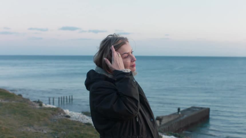 A thoughtful woman in a winter coat enjoys a peaceful, windy moment by the sea before walking out of frame in slow motion