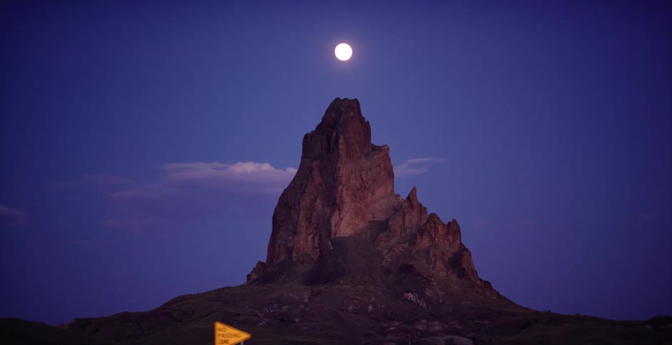 A glowing full moon hangs in the deep blue night sky directly above a majestic desert rock formation