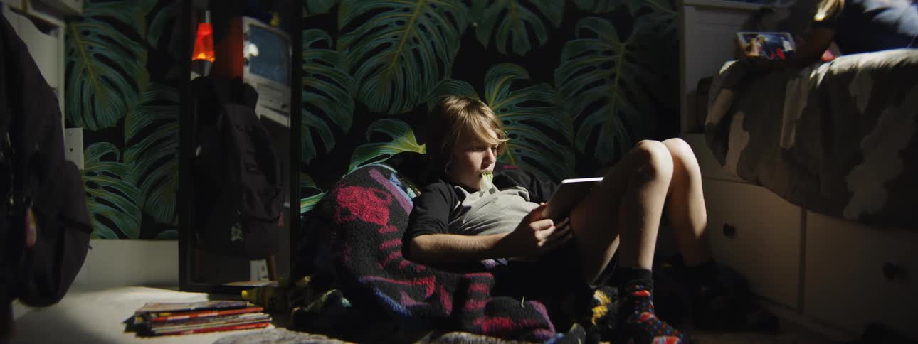 A boy engrossed by a tablet blows a bubblegum that pops while his sister uses a tablet on the bed behind him