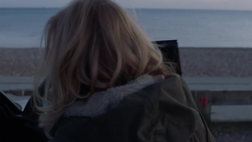 A mature woman walks up to her car, unlocks it with a key fob, and gets into the driver's seat by a pebble beach at dusk