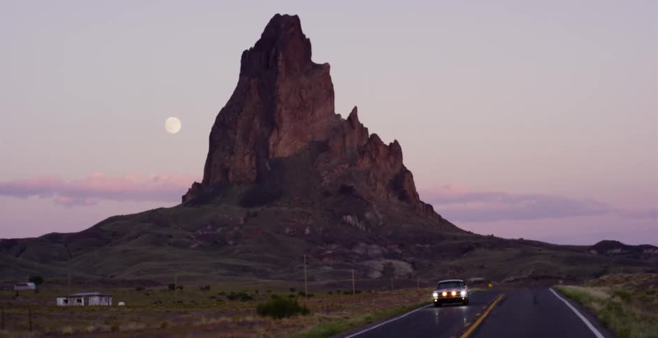 A classic car drives down an empty desert road towards an impressive rock formation as the full moon rises at dusk