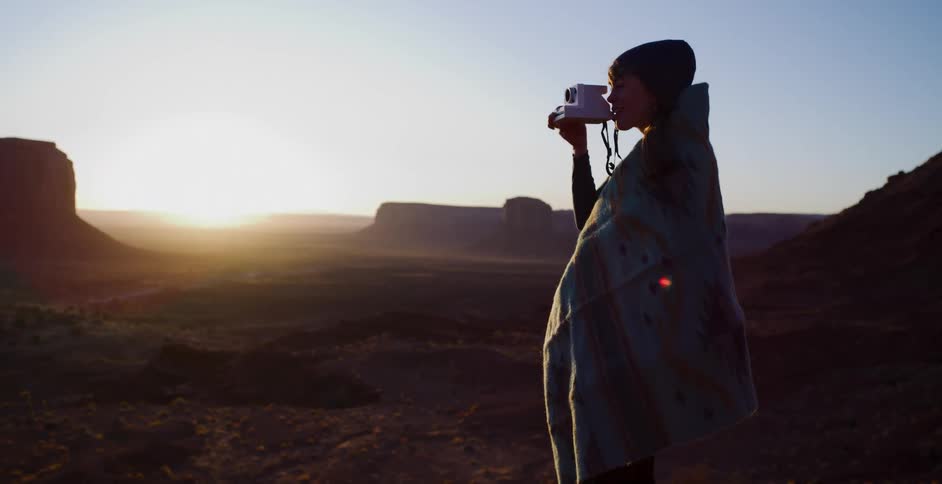 A young woman wrapped in a blanket takes a picture of the majestic desert sunrise with an instant camera in slow motion