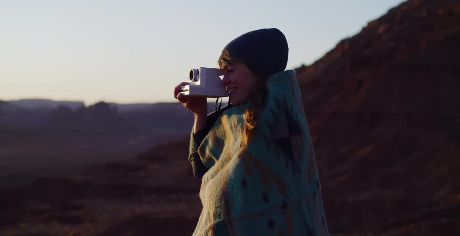 An adventurous woman bundled in a blanket takes a photo of the stunning desert scenery with a vintage instant camera at dawn