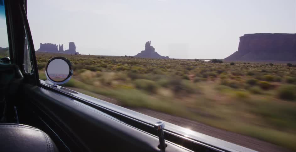 A scenic view of Monument Valley passes by from the window of a classic car driving through the desert