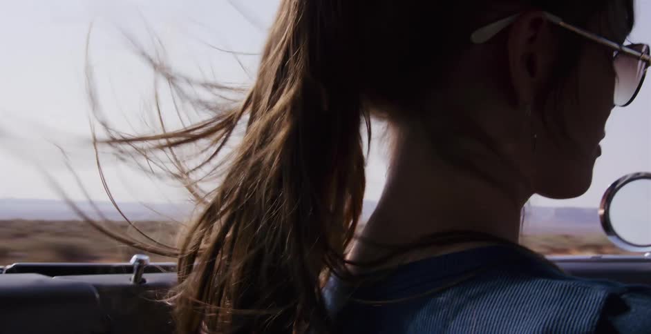 Slow motion shot of a woman's brown ponytail blowing in the wind while she drives a convertible through a desert landscape