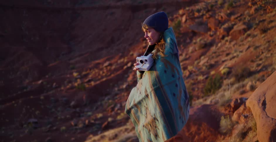 A happy woman wrapped in a warm blanket takes pictures with an instant camera in the desert at sunset