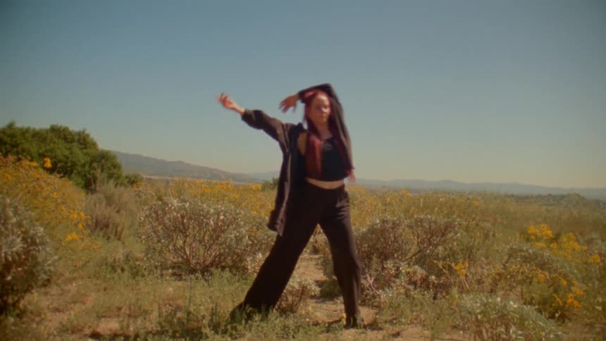 A woman with vibrant red hair dances freely in slow motion amidst a beautiful field of yellow wildflowers on a sunny day