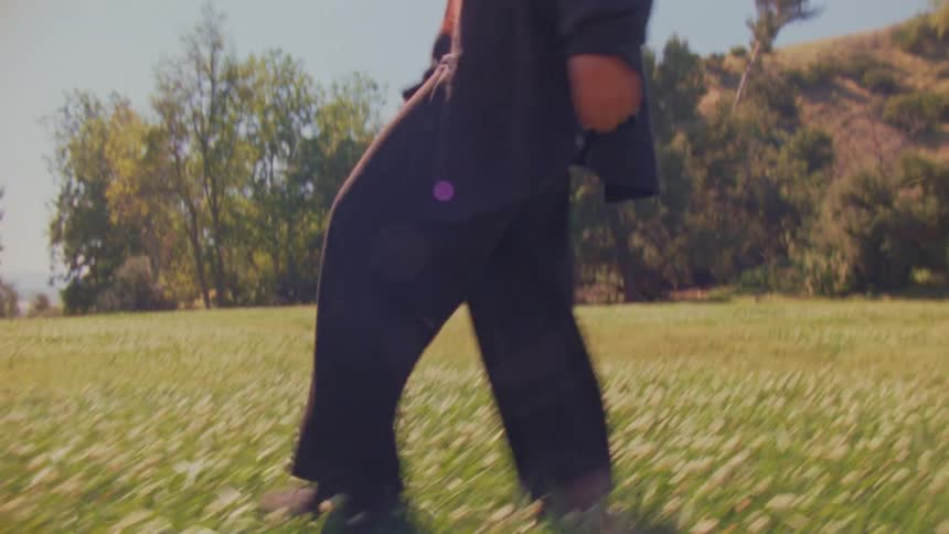 A mystical woman with red hair walks slowly through a sun-drenched meadow in a flowing black cloak
