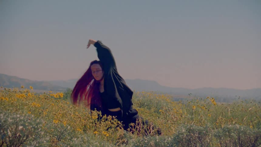A woman with red hair performs an expressive slow-motion dance in a sunny field of yellow wildflowers