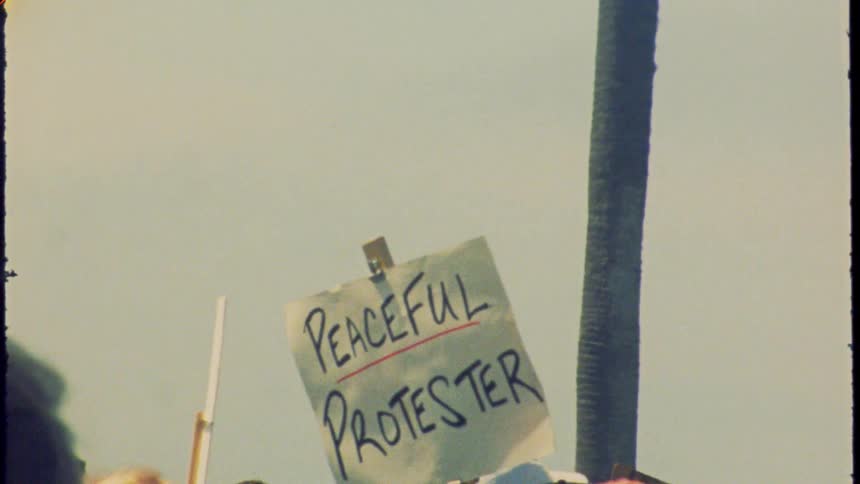 A 'Peaceful Protester' sign is held high in a crowd during a demonstration, captured on vintage film