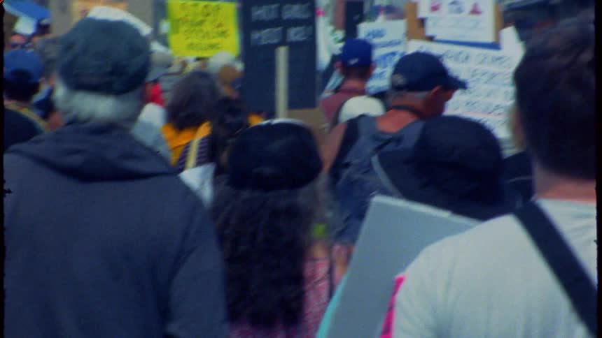 A handheld, vintage-style shot follows a crowd of political protestors as they march through a city street with signs and an American flag