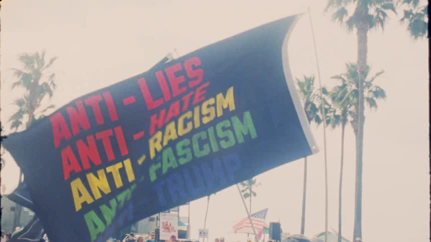 Vintage-style slow-motion footage shows large 'Resist' and anti-racism banners waving in the wind at a political protest with palm trees in the background