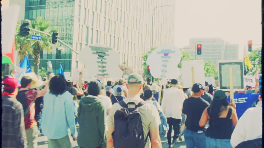 Vintage film-style footage from behind a crowd of protestors marching down a city street with signs