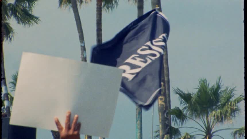 A 'RESIST' flag and blank protest sign wave in slow motion against palm trees in vintage Super 8 footage