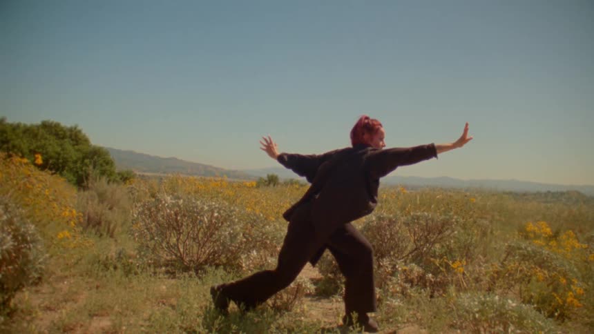 A woman with red hair performs a slow, expressive dance in a sun-drenched field of yellow wildflowers