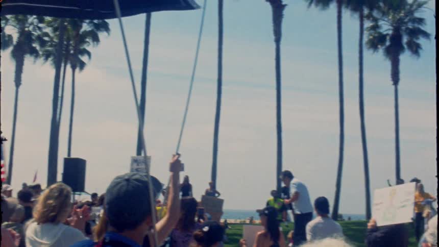 Vintage film captures a political protest on a sunny beach, with American and anti-fascist flags waving over the crowd