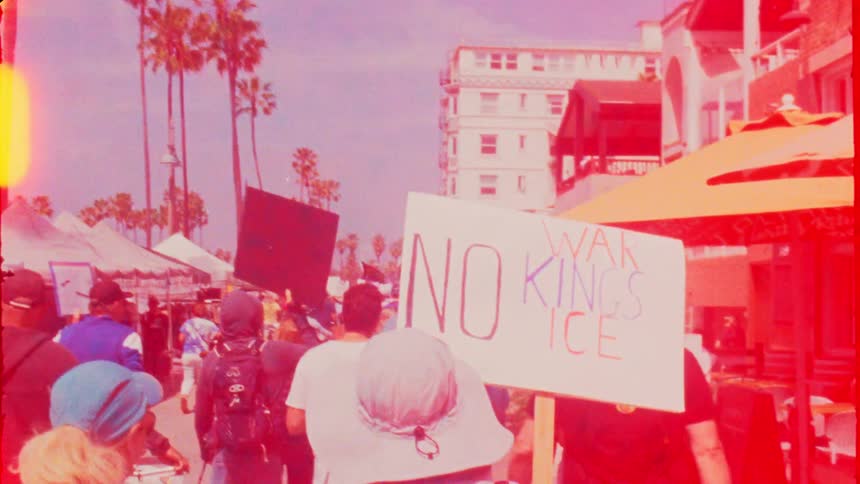Retro film-style footage follows protestors marching with signs down a sunny, palm-lined street