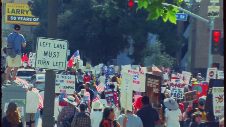 Vintage film footage captures a large, diverse crowd of people marching in a street protest with various political signs