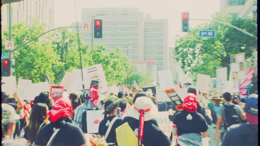 Vintage 8mm film style footage follows a crowd of protestors marching down a sunny city street with signs and banners