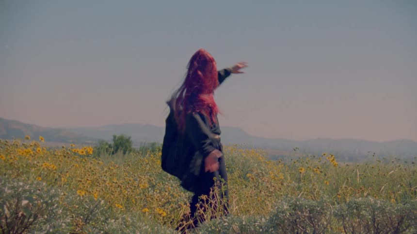 A free-spirited woman with vibrant red hair dances passionately in a sun-drenched field of yellow wildflowers in slow motion